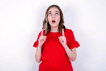 Sticker - young caucasian woman wearing red T-shirt over white background  amazed and surprised looking up and pointing with fingers and raised arms.