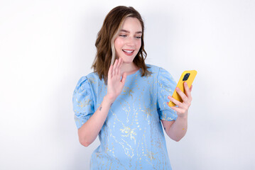 Portrait of happy friendly young caucasian woman wearing blue T-shirt over white background taking selfie and waving hand, communicating on video call, online chatting.