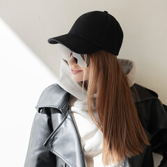 Wall Mural - Fashionable hipster beautiful woman with trendy sunglasses with black mock-up cap in fashion casual outfit with hoodie and leather jacket stands near a beige wall with sunlight. Female street clothes