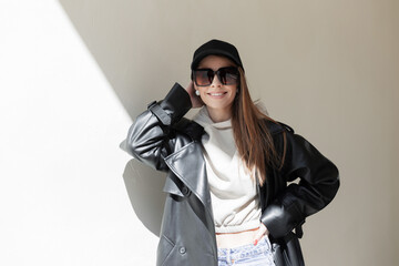 Sticker - Happy beautiful young fashion happy hipster woman with a smile with sunglasses and a cap in a fashionable leather coat and hoodie standing near the wall in the sunlight