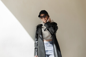 Wall Mural - Trendy fashionable happy beautiful young girl with a smile with cool sunglasses and a black cap mockup in a fashion leather coat with a hoodie and jeans stands near the wall in the street with light