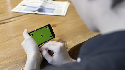 Poster - Smartphone horizontal orientation with a green screen: shot over the shoulder of a young man. Stock Footage. Man sitting at the table, tapping and scrolling on the chromakey of the phone.