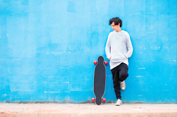 Portrait of a young man with a longboard at his side and blue wall in the background.