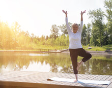 Full-length Portrait Of A Caucasian Elderly Woman In Yoga Vrikshasana Poses, Tree Pose, Against The Background Of A Lake In A Park. Health And Active Lifestyle Of The Elderly.