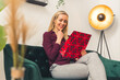 Beautiful smiling blondie sitting on the sofe and with amusement looking at her present - room background. High quality photo