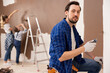 © ABCreative - A smiling brunette man paints the wall in brown color with a small roller. Behind him, from the back, are his wife and daughter. They are all involved in repairs.