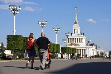 Wall Mural - Couple with baby pram walking in VDNKh park on people background. Soviet architecture, russian attractions in summer