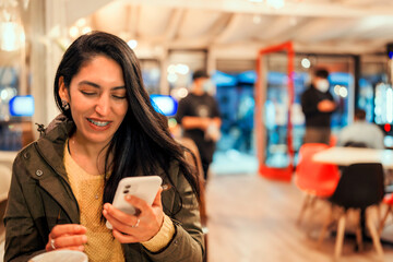 young latin woman holding a cell phone in her hands sitting and smiling in a cafe 