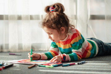 A cute little girl sits on the floor in the rays of the sun and draws a rainbow with colorful markers. She is dressed in a colored jumper. Creation. Childhood.