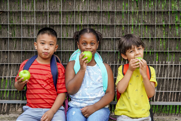 multiethnic children with backpacks eating apples at school entrance. daily rate of fruit.