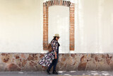 Fototapeta Na drzwi - Single latin adult woman with sunglasses and hat walks through the colonial style streets with cobbled white wall, relaxed she strolls as a tourist on vacation or weekend
