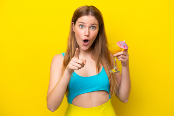 Wall Mural - Young caucasian woman holding a cocktail isolated on yellow background surprised and pointing front