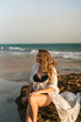 Chica joven de pelo largo sentada en una roca a la orilla del mar posando y mirando el atardecer