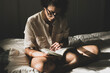 Young woman reading in her room with the light from the window sitting on the bed. Woman spending her leisure time reading book at night before sleeping. Girl enjoying her reading time
