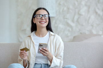 Young beautiful woman in glasses smiling and happy doing online shopping in online store sitting on sofa at home, holding mobile smartphone and bank credit card