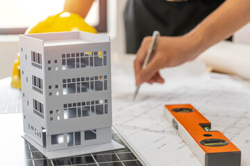 Wall Mural - building model on workplace desk with architect or construction worker working with blueprint on desk in office center at construction site, industrial, construction contract and contractor concept