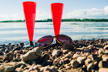 In The Summer, Two Red Glasses Of Sparkling Champagne Or Prosecco In The Bay And Sunglasses On A Pebbly Beach