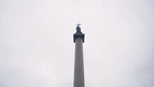 Alexandrian Pillar In Saint Petersburg. Action. Bottom View Of Architectural Monument In Form Of Pillar With Angel Holding Cross On Top. Alexander Column On Background Cloudy Sky
