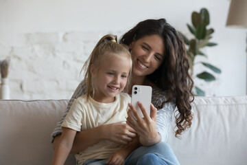 Wall Mural - Happy mom holding cute little daughter kid in arms, using smartphone on couch at home, talking on video call, taking selfie. Blogger mother hugging child with love, tenderness, cellphone camera