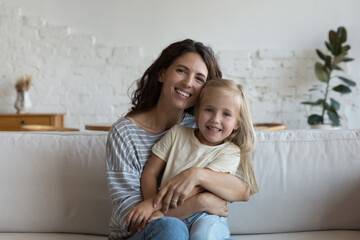 Wall Mural - Happy joyful mom hugging happy sweet little preschool daughter, holding kid in arms, sitting on couch at home, looking at camera, smiling. Motherhood, family concept. Video call head shot portrait