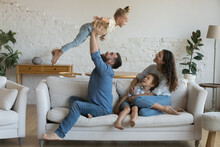 Happy Young Parents Having Fun With Little Kids, Enjoying Family Leisure, Activity On Couch At Home. Playful Daddy Lifting Joyful Daughter Girl Up In Air, Mom Hugging Son. Family Activity