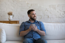 Happy Dreamy Cellphone User Man Holding Mobile Phone, Relaxing On Sofa, Looking Away, Smiling At Good Thoughts, Dreaming, Thinking Over Good News, Message, Chatting Online. Communication Concept