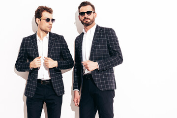 Portrait of two handsome confident stylish hipster lambersexual models. Sexy modern men dressed in black same elegant suit. Fashion male posing in studio near white wall. In sunglasses