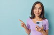 © WHstudio Leushin N - Beautiful Asian woman has dark hair holds mobile phone chats online browses social networks learn foreign language via special app wears purple t shirt points away on blank space isolated on blue wall