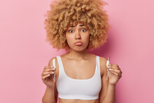 Horizontal Shot Of Pretty Displeased Young Woman With Curly Hair Holds Two Tampons Chooses Best Absorbency Product Dressed In White Top Looks Sadly Isolated Over Pink Background. Menstruation