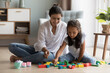 © fizkes - Cheerful pretty Indian mother and little daughter kid playing on heating floor at home, arranging colorful toy blocks. Nanny, daycare teacher entertaining child, enjoying learning game