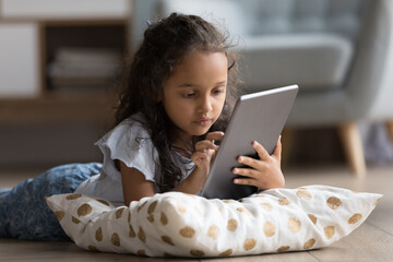 Focused gen Z preteen school kid resting on heating floor with soft pillow, using learning app on digital tablet computer, reading book online, doing homework, browsing Internet, social media
