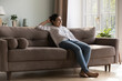 © fizkes - Cheerful carefree young Indian woman enjoying relaxation co comfortable sofa, looking at window away, dreaming, thinking over good news, plans. Home leisure concept. Full length