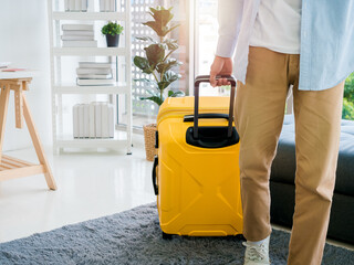  A man with yellow luggage. A male in denim shirt and brown trousers walking while holding a suitcase at the handle in office room at home. Summer holiday travel vacation concept. Time to journey.