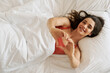Top view of beautiful young woman gesturing heart shape and smiling while lying in bed