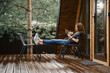 © boomeart - Young woman sits on terrace of a-frame house with mobile phone