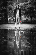 Young Man In The Black Cardigan With The Red Rose In The Hand Stands In Front Of The Puddle With The Reflection Of Himself. The Behind The Mirror Man. Sad Romance Concept.