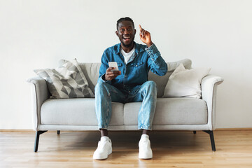 Wall Mural - Excited African American Guy Holding Smartphone Having Idea Sitting Indoors