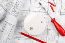 Smoke Detector Or Fire Alarm Sensor With Rolls Of Architectural Drawing Background With Tools And Screws, House Safety Or Security Concept, Flat Lay Top View