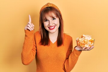 Sticker - Redhead young woman holding potato chips smiling with an idea or question pointing finger with happy face, number one