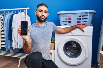 Wall Mural - Middle east man with beard showing smartphone screen and washing machine looking at the camera blowing a kiss being lovely and sexy. love expression.