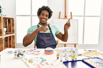 Wall Mural - Young african american man sitting on the table at art studio smiling and looking at the camera pointing with two hands and fingers to the side.