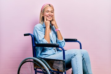 Wall Mural - Beautiful blonde woman sitting on wheelchair smiling with open mouth, fingers pointing and forcing cheerful smile