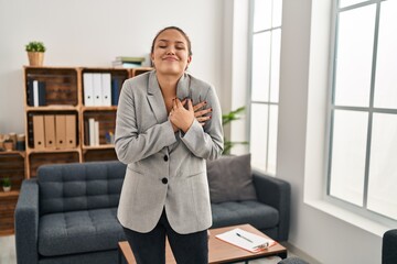 Wall Mural - Young hispanic woman working at consultation office smiling with hands on chest, eyes closed with grateful gesture on face. health concept.