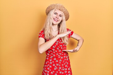 Poster - Beautiful caucasian woman with blond hair wearing summer hat gesturing with hands showing big and large size sign, measure symbol. smiling looking at the camera. measuring concept.