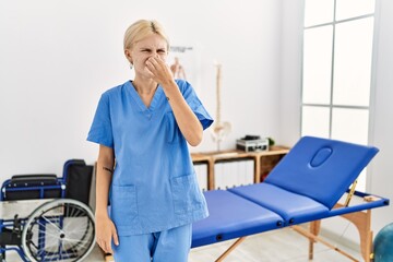 Sticker - Beautiful caucasian physiotherapist woman working at pain recovery clinic smelling something stinky and disgusting, intolerable smell, holding breath with fingers on nose. bad smell