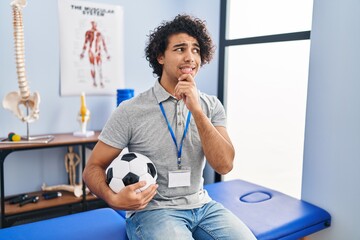 Canvas Print - Hispanic man with curly hair working as football physiotherapist thinking worried about a question, concerned and nervous with hand on chin