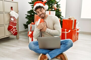 Sticker - Young hispanic man smiling happy holding christmas gift using laptop at home.