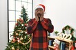 African american man wearing santa claus hat standing by christmas tree afraid and shocked, surprise and amazed expression with hands on face