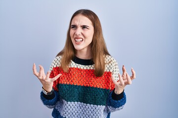 Poster - Young hispanic girl standing over blue background crazy and mad shouting and yelling with aggressive expression and arms raised. frustration concept.