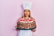 Beautiful hispanic woman wearing cook uniform and hat and birthday cake making fish face with mouth and squinting eyes, crazy and comical.